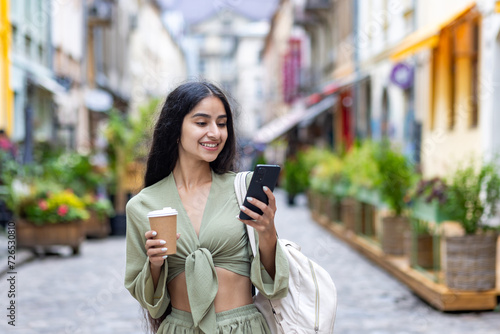 Photography Indian young smiling girl walking in the city, holding a cup of coffee and using a mobile phone