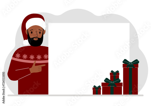 A man in a Christmas or New Year's sweater with a red sana cap points his finger at a banner, a blank poster or a large white sheet of paper with space for text.