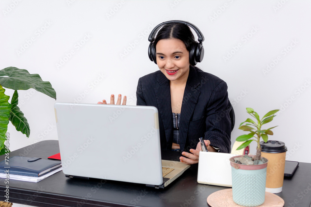 Cheerful female office employee using a laptop while working at her desk with headphones, coffee, and plants. Discussing via video call to a client. Cheerful female office employee using a laptop while working at her desk with headphones, coffee, and plants. Discussing via video call to a client.