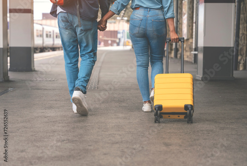 Happy loving couple with luggage and backpack walking along railway platform as they are ready to travel on holiday. Man and woman holding hands, looking back and smiling. Travel by train concept