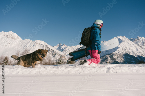 Female snowboarder freerider  hiking with dog on ski resort, winter sport outdoor, sunny day in mountains