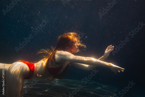 Young redheaded woman swimming underwater in a pool.