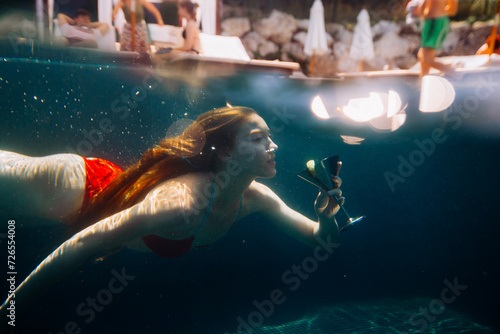 Fotografie A beautiful red-haired woman poses with a cocktail under water.
