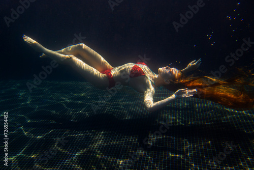 A beautiful sporty girl poses underwater with loose hair against the bright rays of the sun from the surface.