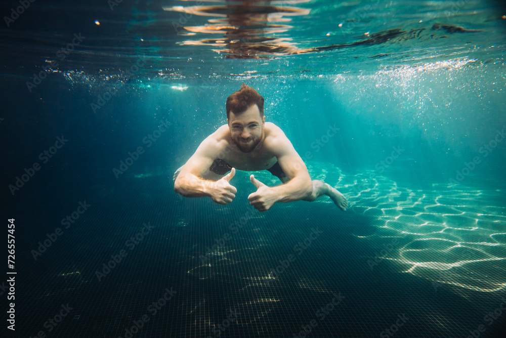 Young man shows thumbs up underwater in swimming pool. Summer vacation concept.