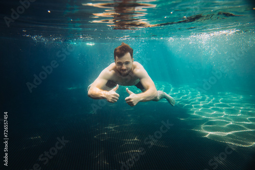 Young man shows thumbs up underwater in swimming pool. Summer vacation concept.