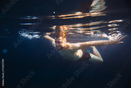 A handsome young man swims underwater in a swimming pool while on vacation.