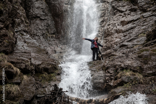 A girl and a dog stand against the backdrop of a waterfall. Beautiful nature. Splashes of water. American Staffordshire Terrier. Mountains.