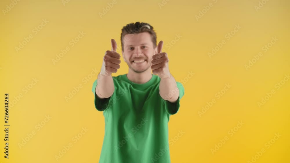 A young man stands on a yellow background and raises his hands showing thumbs up. A smiling guy in a green t-shirt shows thumbs up to the camera in approval. A young man stands on a yellow background and raises his hands showing thumbs up. A smiling guy in a green t-shirt shows thumbs up to the camera in approval.