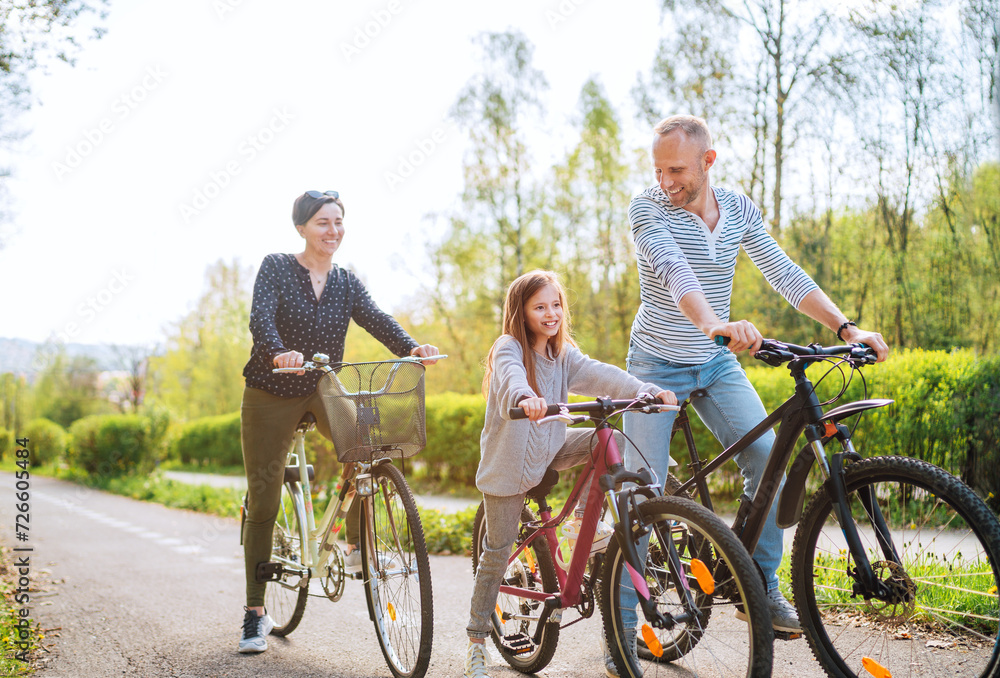 Obraz premium Smiling father and mother with daughter during summer outdoor bicycle riding. They enjoy togetherness in the summer city park. Happy parenthood and childhood or active sport life concept image.