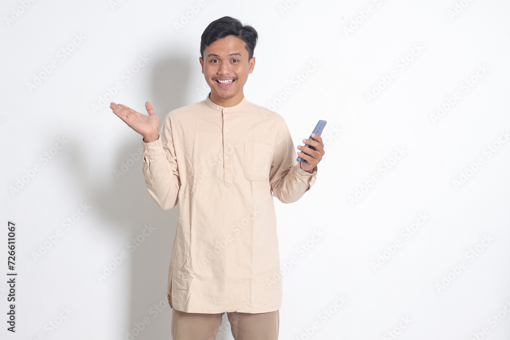 Portrait of young excited Asian muslim man in koko shirt holding mobile phone, pointing and showing product with finger. Social media concept. Isolated image on white background Portrait of young excited Asian muslim man in koko shirt holding mobile phone, pointing and showing product with finger. Social media concept. Isolated image on white background