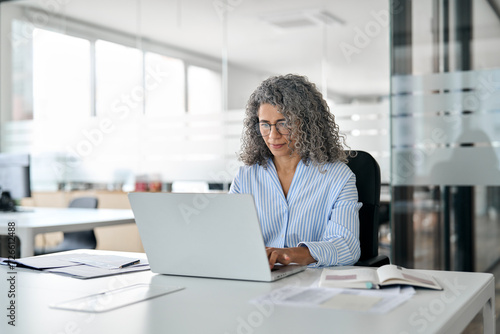 Serious busy mature middle aged professional business woman, older lady manager executive leader wearing glasses looking at laptop using computer in office working on digital project sitting at desk.