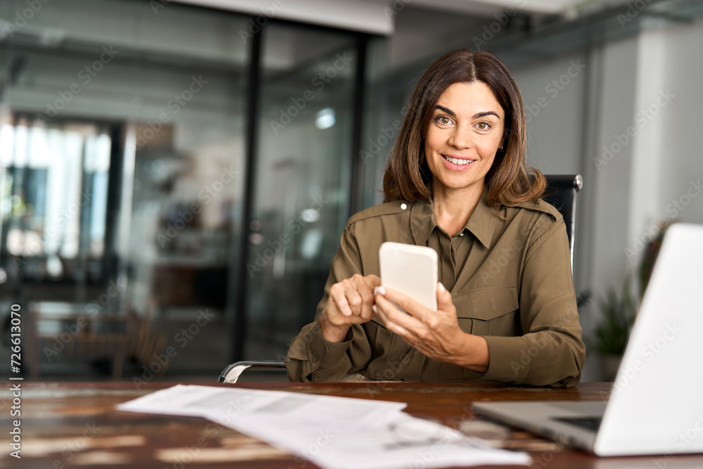 Smiling mature business woman executive using mobile phone working in office. Happy middle aged businesswoman holding smartphone, professional female executive looking camera with cellphone. Portrait. Smiling mature business woman executive using mobile phone working in office. Happy middle aged businesswoman holding smartphone, professional female executive looking camera with cellphone. Portrait.