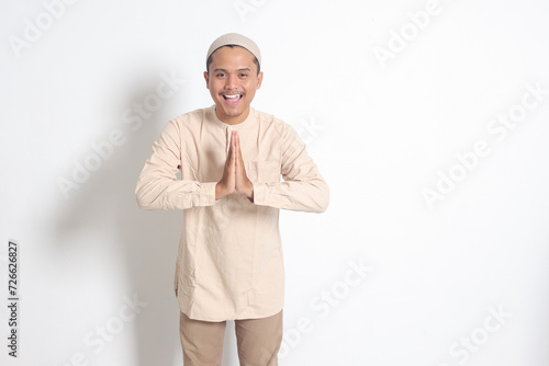 Portrait of attractive Asian muslim man in koko shirt with skullcap showing apologize and welcome hand gesture. Apology during eid mubarak. Isolated image on white background
