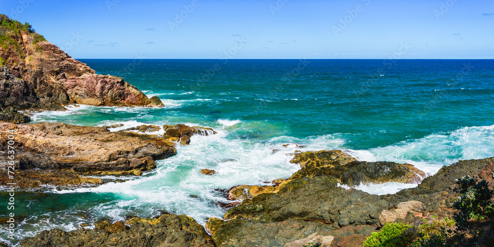 Australian coast, view from a cliff to the blue ocean with a rocky shore on a sunny day. Sea landscape, waves crashing on volcanic rocks at the shore.