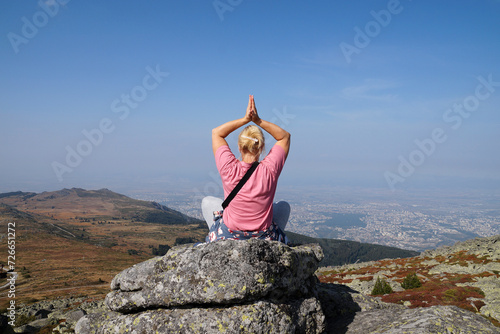 blonde woman meditates in a yoga pose with her arms raised above her head on the top of a mountain
