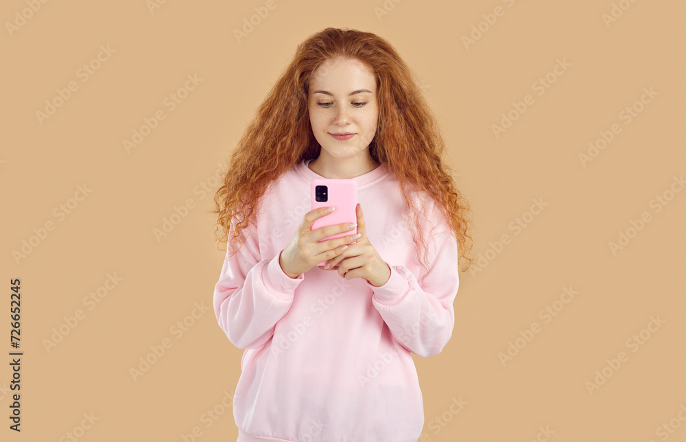 Calm, interested, relaxed redhead pretty curly girl in pink sweatshirt is looking at smartphone screen, reading message or watching video, talking video chat on beige background. Online life concept. Calm, interested, relaxed redhead pretty curly girl in pink sweatshirt is looking at smartphone screen, reading message or watching video, talking video chat on beige background. Online life concept.