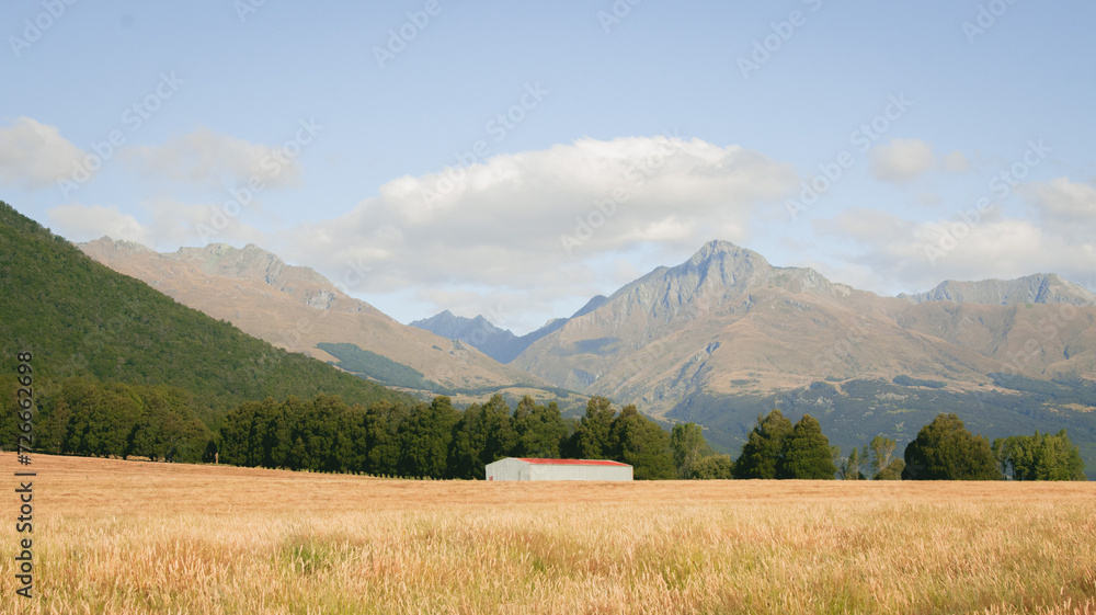 A large red-roofed shed with a mountainous landscape behind it. Surrounded by pine trees. With clouds in the sky