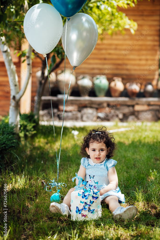 Joyful little toddler girl celebrating her two months in the garden sitting on the lawn with her cake