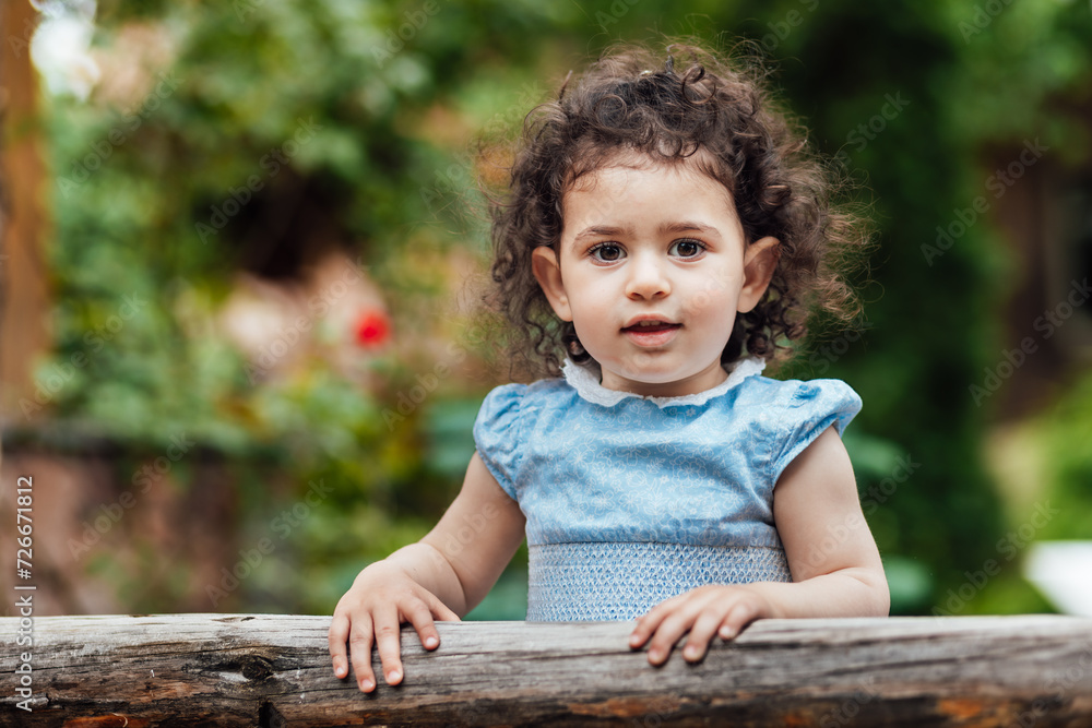 Happy, cheerful toddler girl with curly hair leaning on wooden fence and looking at camera