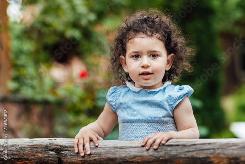 Obraz na płótnie Happy, cheerful toddler girl with curly hair leaning on wooden fence and looking at camera w salonie