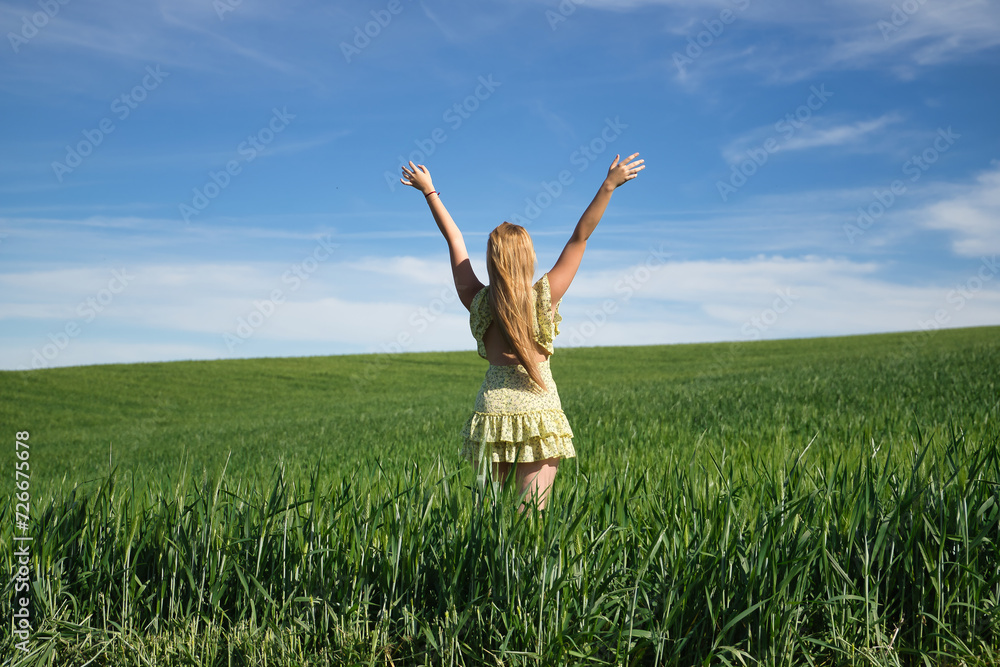 Young, beautiful, blonde woman in a yellow dress, arms raised in the middle of a green wheat field, seen from the back, alone, calm and peaceful. Concept relaxation, tranquility, calm, fields, meadows Young, beautiful, blonde woman in a yellow dress, arms raised in the middle of a green wheat field, seen from the back, alone, calm and peaceful. Concept relaxation, tranquility, calm, fields, meadows