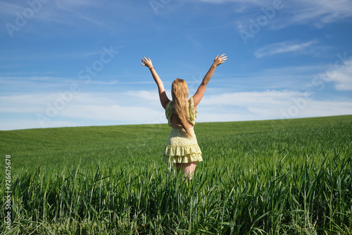 Young, beautiful, blonde woman in a yellow dress, arms raised in the middle of a green wheat field, seen from the back, alone, calm and peaceful. Concept relaxation, tranquility, calm, fields, meadows