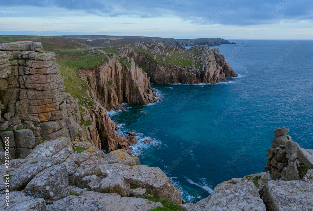 A dramatic landscape photo of a bay in Land＇s End, Cornwall, with the sheer granite cliff face and deep blue water below. 