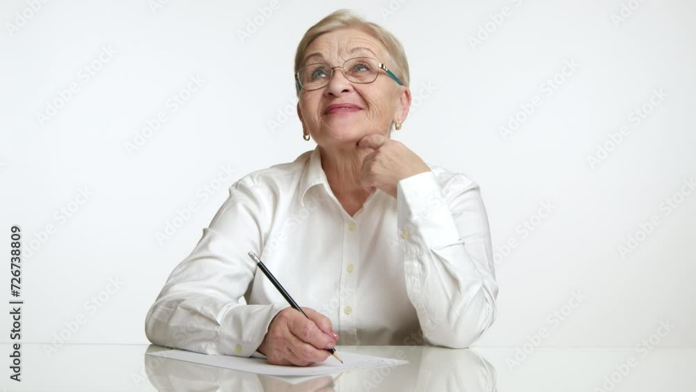 Happy pretty smart elegant senior lady in spectacles, white shirt sitting at table in white studio, dreaming, making notes on sheet of paper with a pencil. High quality 4k footage Happy pretty smart elegant senior lady in spectacles, white shirt sitting at table in white studio, dreaming, making notes on sheet of paper with a pencil. High quality 4k footage