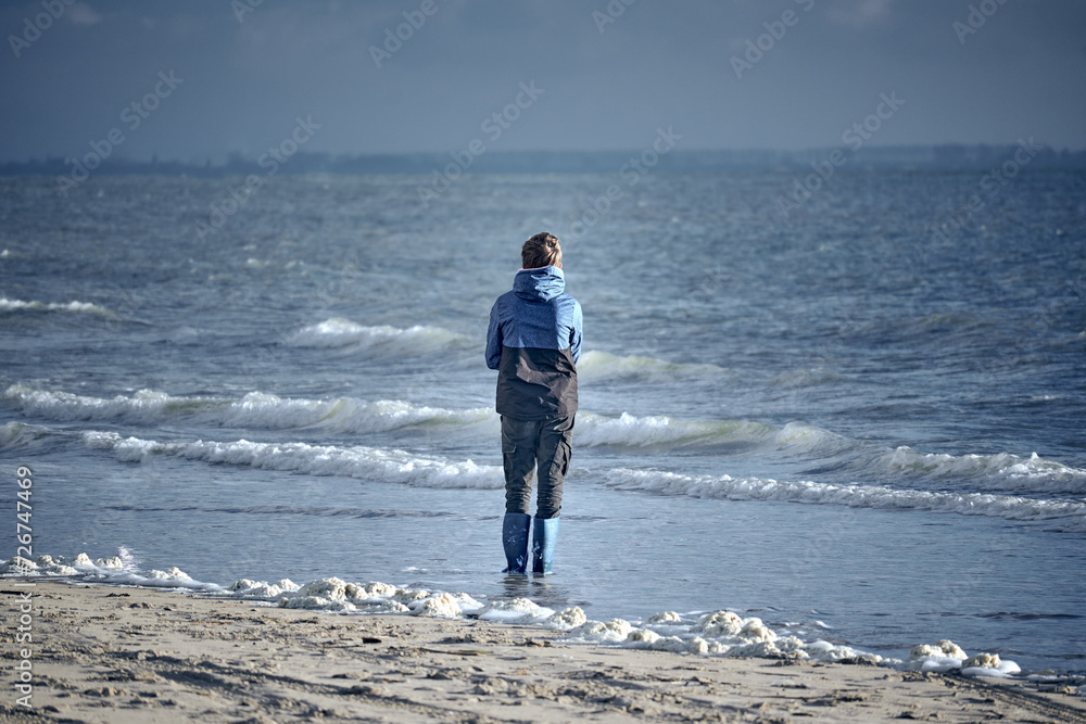 Young boy standing alone on the shore looking out to the sea 