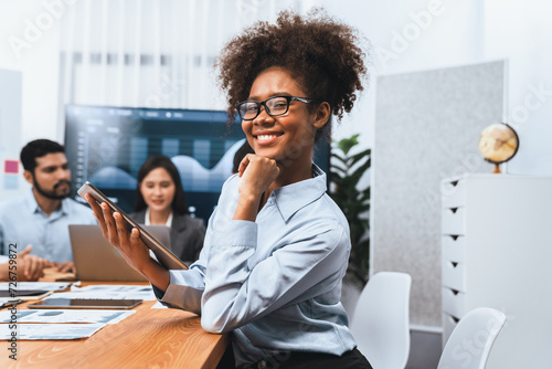 Happy young african businesswoman wearing glasses portrait with group of office worker on meeting with screen display business dashboard in background. Confident office lady at team meeting. Concord photo