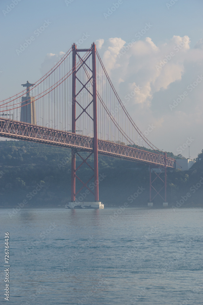 Fototapeta premium Red bridge 25 de Abril Bridge and statue of Cristo Rei during a slightly misty day, Lisbon, Portugal