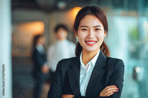 Portrait of a attractive smiling asian businesswoman in a suit standing in a modern business company office  with her workers standing in the blurry background
