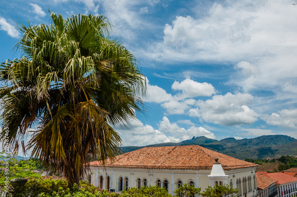 The peak of Itacolomi in the background on a day with clouds and sun and in the foreground a palm tree and an old house in the city of Ouro Preto