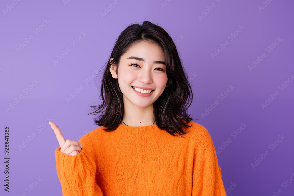 Young Asian woman in orange sweater pointing with a smile on purple background Young Asian woman in orange sweater pointing with a smile on purple background