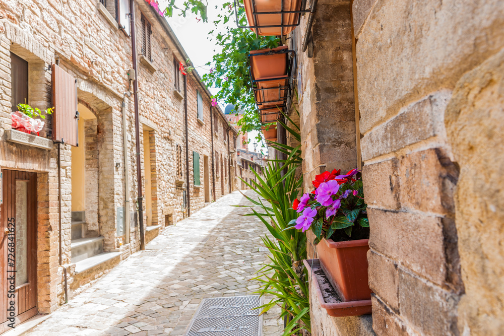 Sun shining on one side of narrow cobblestone street lined by typical European stone block residential with colourful potted flowers.