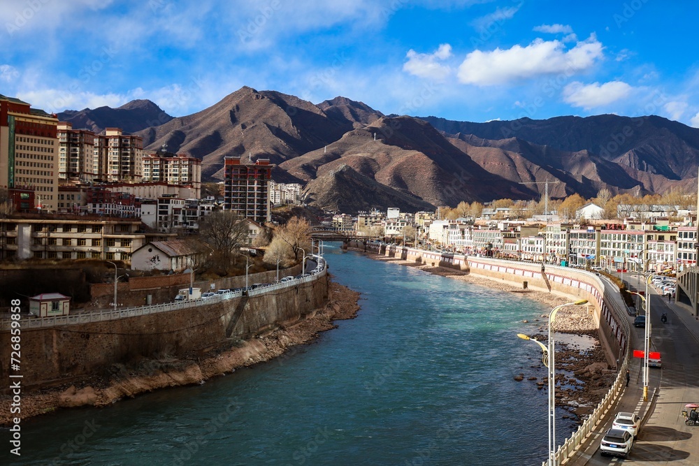 Scenic view of the upper stream of the Mekong River in Tibet, Qamdo, flanked by towering highrise buildings, capturing the juxtaposition of nature and urban development in this picturesque landscape.