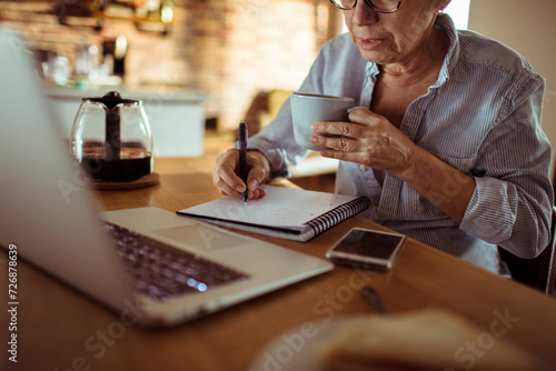 Senior woman using a laptop and writing in a notepad at home