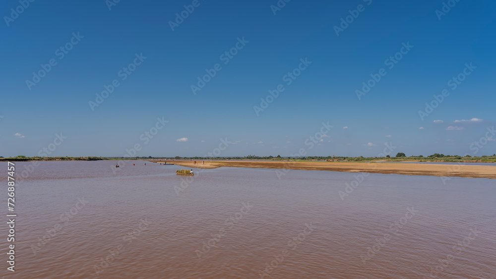 A wide calm river with red-brown water. A man in a boat loaded with hay is floating in a riverbed. Tiny figures of people washing clothes are visible. Clear blue sky. Copy space. Madagascar.