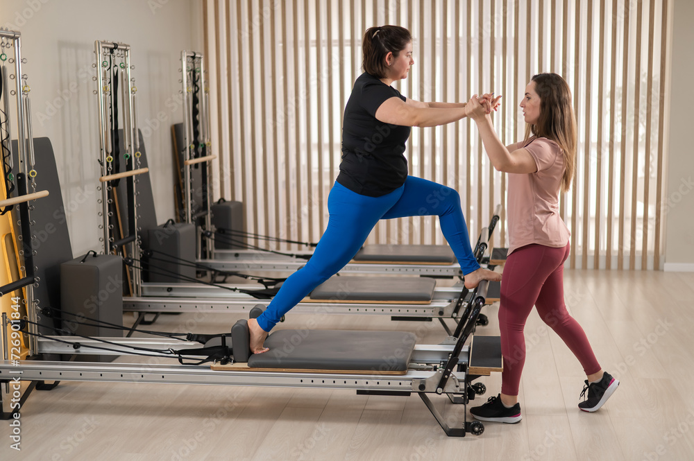 Overweight caucasian woman doing pilates exercises on reformer with personal trainer. Overweight caucasian woman doing pilates exercises on reformer with personal trainer.