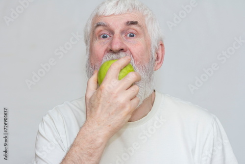 an elderly gray-haired man with a beard holds a large green apple in his hands and eats it