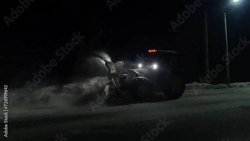 snow removal equipment in the form of a tractor operating at night and throwing snow off the roadway into a ditch photo
