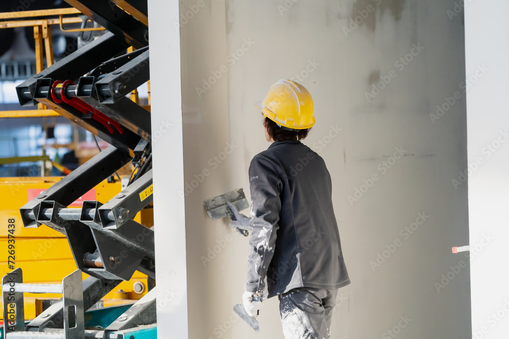 Fototapeta premium Female construction worker applies plaster on construction project wall with construction lift equipment background