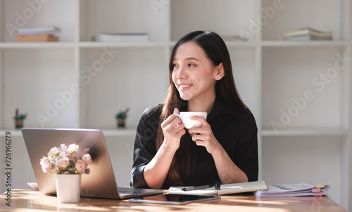 Smiling businesswoman happily sips coffee while working on her laptop in a cozy office setting