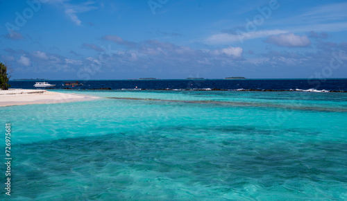 View from lagoon to the ocean. On the left side is the coast of the island with a sandy beach and two small boats. On the horizon are small islands with palm trees. There are some clouds in the sky. © Denis