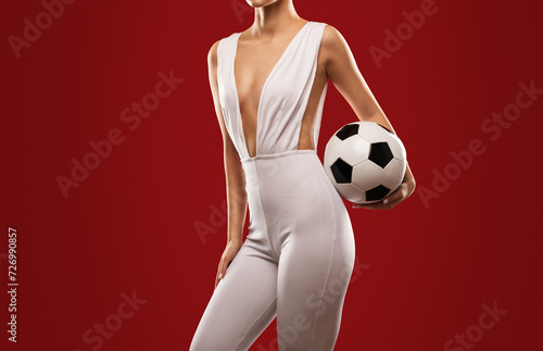 A beautiful sexy girl in a white jumpsuit holds a black and white soccer ball on a red background.