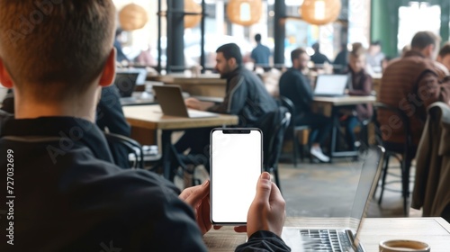 In a bustling city cafe, a young professional sits with the white screen smartphone mockup, showcasing a sleek design against the backdrop of people working on laptops, mockup