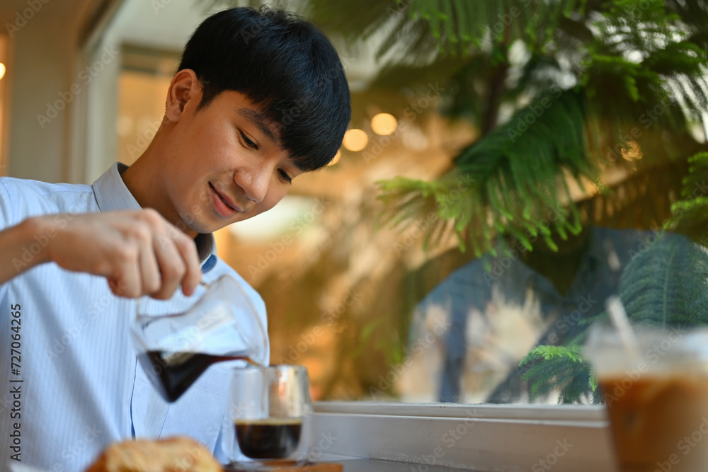 Fototapeta premium A Handsome Young Asian male pouring a black coffee into the glass in the cafe