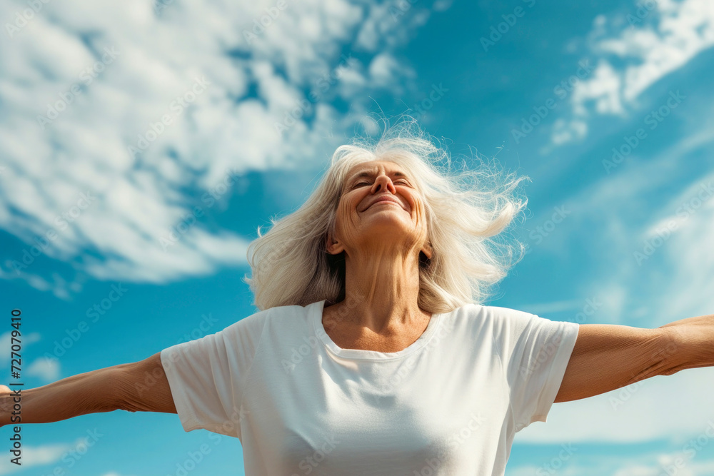Joyful ederly woman with white a t'shirt enjoying freedom standing with open arms and a happy smile looking up towards the sky