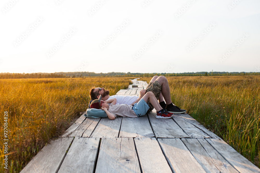 Father and son relaxing on wooden boardwalk through swamp. Man with child resting in wildlife national park at sunset. Concept of ecotourism, hiking, vacation, travel with children. Selective focus.